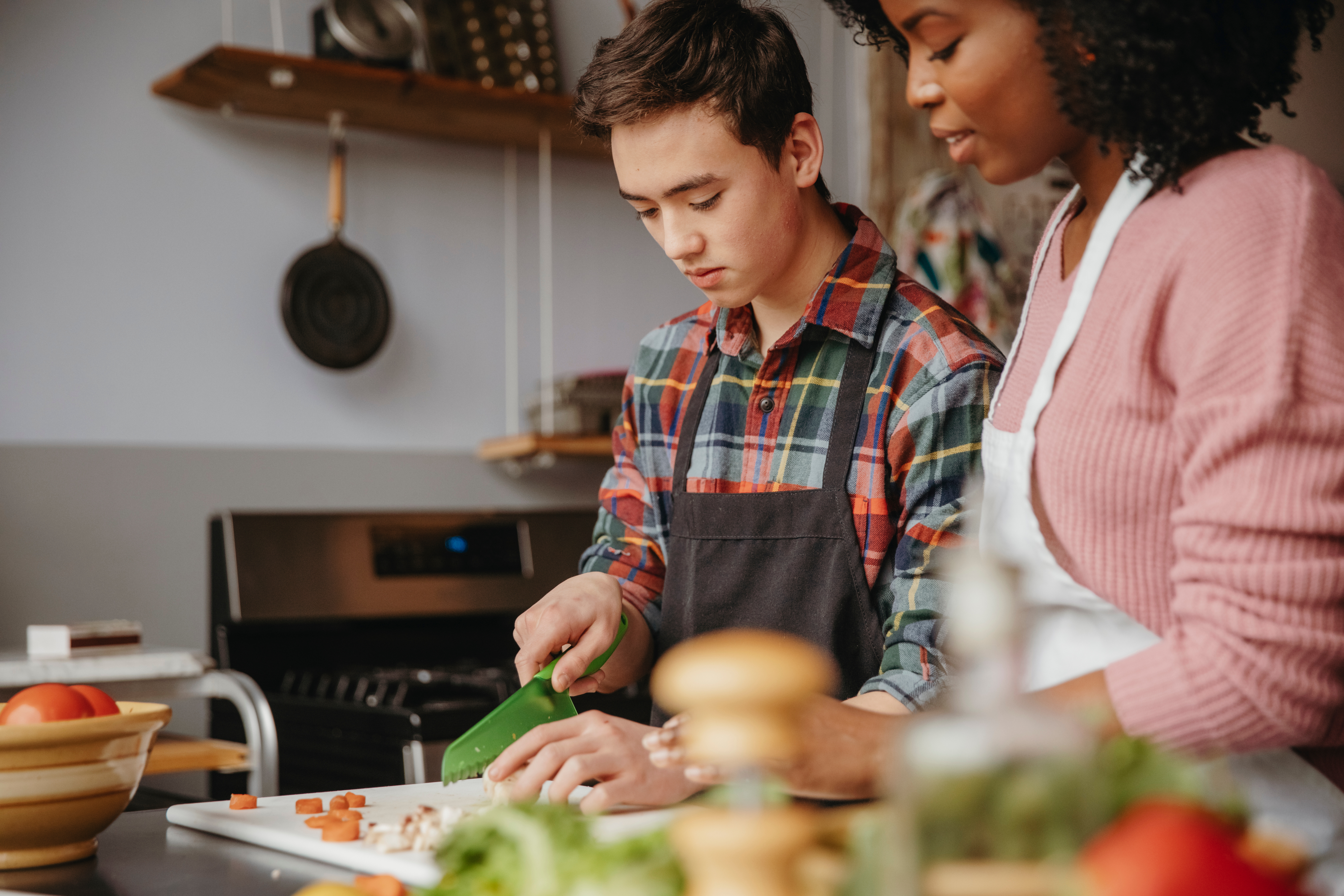 Female chef assisting student in chopping vegetables on table at cooking class female chef with curly hair in a pink jumper chopping vegetables next to a young boy in a checkered shirt wearing a black apron